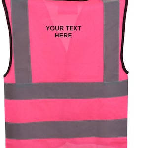 Może przedstawiać: R&oacute;żowa kamizelka bezpieczeństwa z odblaskowymi srebrnymi paskami. Kamizelka ma czarną lam&oacute;wkę i napis "YOUR TEXT HERE" nadrukowany na plecach.