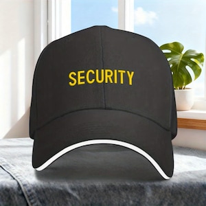 May include: Black baseball cap with the word "SECURITY" embroidered in yellow. The cap has a white trim along the brim. The hat is sitting on a gray surface.