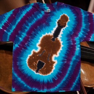Peut inclure: Un t-shirt tie-dye avec une silhouette de violon marron et noir au centre. Le t-shirt a un motif en spirale bleu, violet et blanc.
