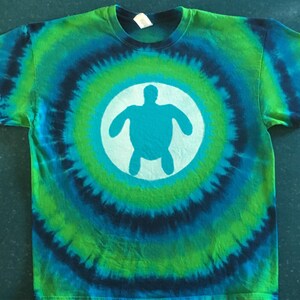 May include: A tie-dye t-shirt with a green, blue, and black swirl pattern. A white circle with a blue silhouette of a sea turtle is centered on the shirt.
