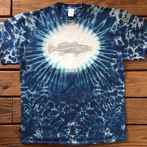 Puede incluir: Una camiseta azul tie-dye con un círculo blanco en el centro que presenta una silueta gris de un pez. La camiseta tiene un patrón circular de azul y blanco.
