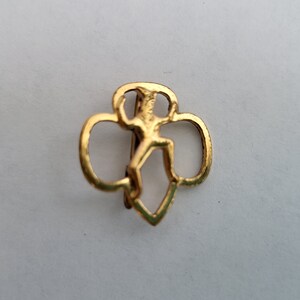 May include: Gold-colored Girl Scout pin featuring a stylized figure of a girl in a running pose, surrounded by three overlapping circles.