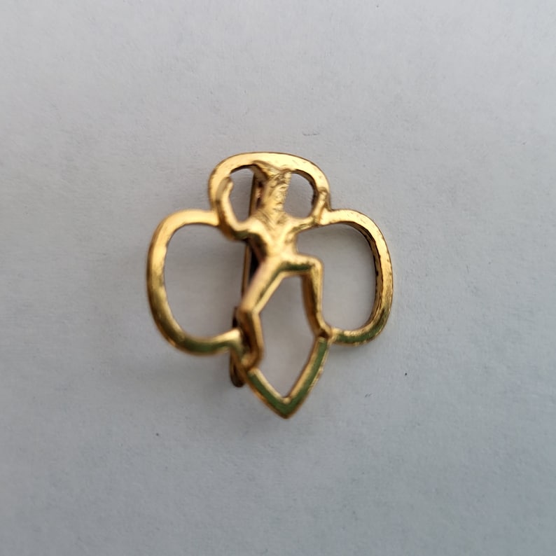 May include: Gold-colored Girl Scout pin featuring a stylized figure of a girl in a running pose, surrounded by three overlapping circles.