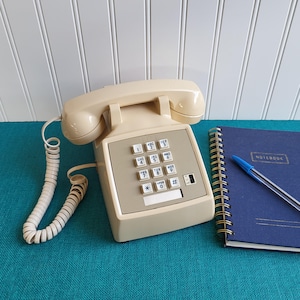 May include: A vintage beige telephone with a push-button keypad and a coiled handset cord. A blue spiral-bound notebook with a pen and the word "NOTEBOOK" is next to the phone. The phone is on a teal surface.