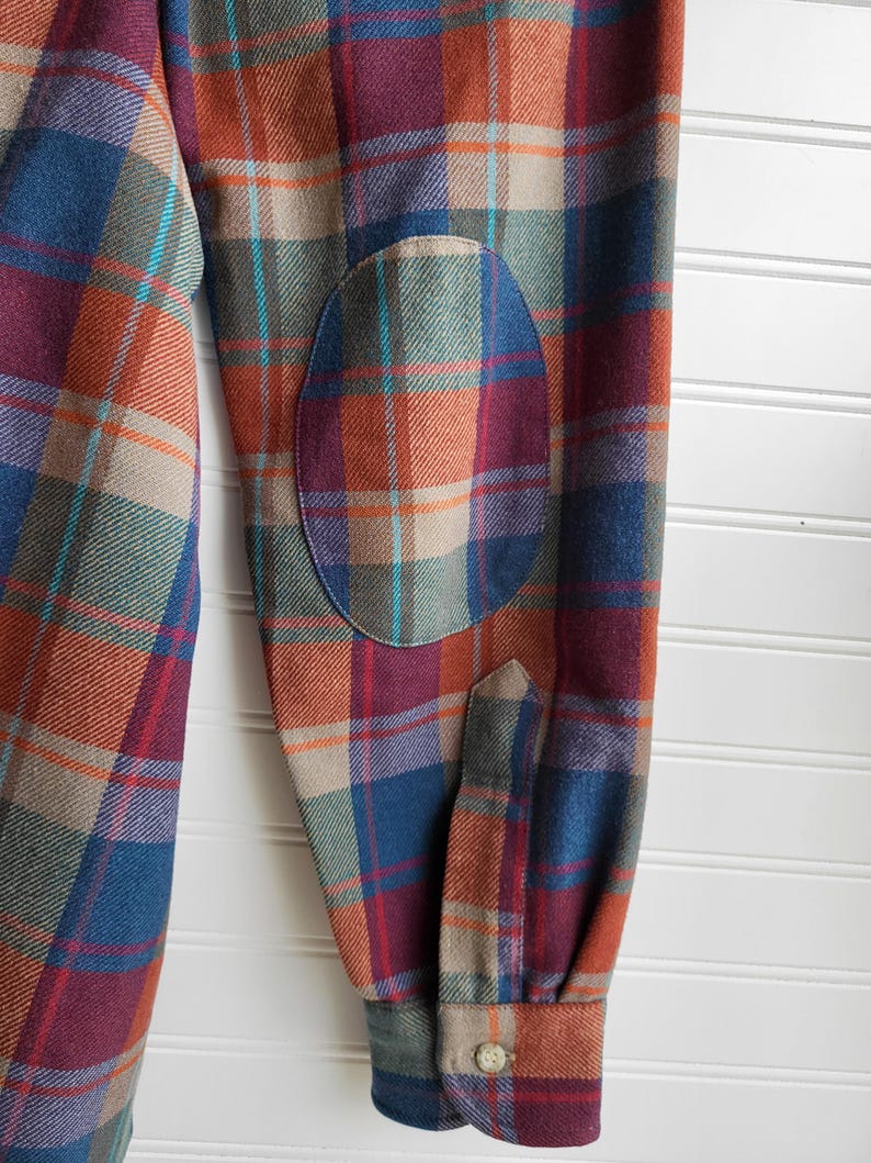 Peut inclure: Gros plan d'une chemise &agrave; carreaux &agrave; manches longues dans les tons bordeaux, bleu, orange et vert. Le coude est assorti. Le poignet a une fermeture &agrave; bouton.