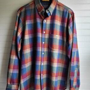 Peut inclure: Une chemise &agrave; carreaux boutonn&eacute;e dans les tons de bleu, orange, rouge et gris, suspendue &agrave; un cintre en bois. La chemise &agrave; manches longues est dot&eacute;e d'un col boutonn&eacute;, d'une poche poitrine et de coudi&egrave;res. La chemise est en tissu doux et tiss&eacute;.