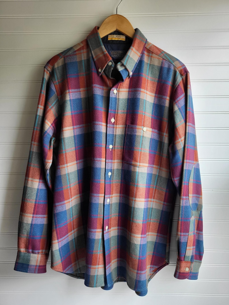 Peut inclure: Une chemise &agrave; carreaux boutonn&eacute;e dans les tons de bleu, orange, rouge et gris, suspendue &agrave; un cintre en bois. La chemise &agrave; manches longues est dot&eacute;e d'un col boutonn&eacute;, d'une poche poitrine et de coudi&egrave;res. La chemise est en tissu doux et tiss&eacute;.