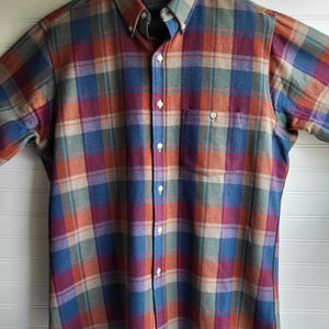 Peut inclure: Une chemise &agrave; carreaux boutonn&eacute;e dans les tons de bleu, orange, violet et sarcelle. La chemise a une poche poitrine et est suspendue &agrave; un cintre en bois. La chemise est en tissu doux et tiss&eacute;.