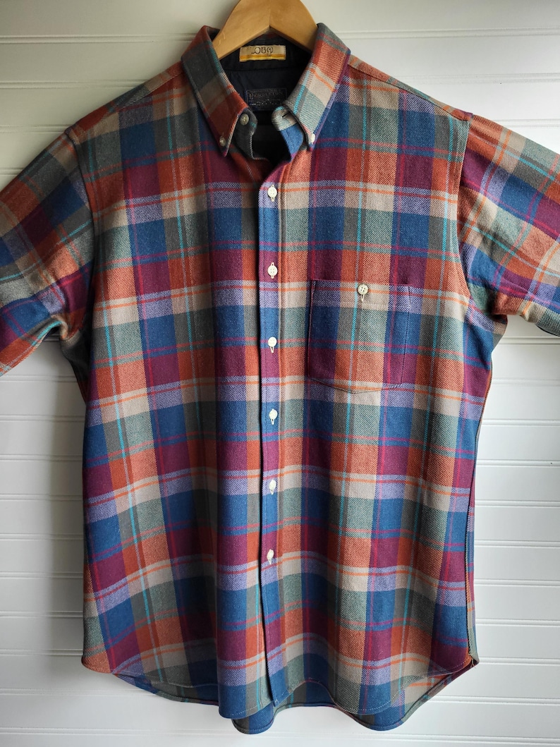 Peut inclure: Une chemise &agrave; carreaux boutonn&eacute;e dans les tons de bleu, orange, violet et sarcelle. La chemise a une poche poitrine et est suspendue &agrave; un cintre en bois. La chemise est en tissu doux et tiss&eacute;.