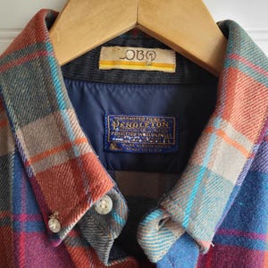Peut inclure: Gros plan d'une chemise &agrave; carreaux Pendleton aux tons de rouge, bleu et gris. La chemise est suspendue &agrave; un cintre en bois. Le col et la doublure int&eacute;rieure sont visibles, avec une &eacute;tiquette "Pendleton".