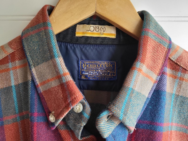 Peut inclure: Gros plan d'une chemise &agrave; carreaux Pendleton aux tons de rouge, bleu et gris. La chemise est suspendue &agrave; un cintre en bois. Le col et la doublure int&eacute;rieure sont visibles, avec une &eacute;tiquette "Pendleton".