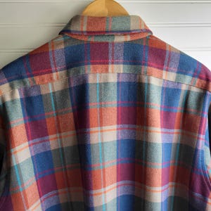 Peut inclure: Une chemise en flanelle &agrave; carreaux multicolores, dans les tons de bleu, orange, rouge et beige, suspendue &agrave; un cintre en bois. Le dos de la chemise est visible, mettant en valeur le motif &agrave; carreaux classique. Le col et les &eacute;paules de la chemise sont mis en &eacute;vidence.