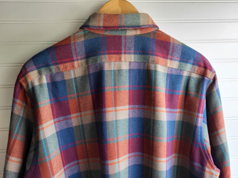 Peut inclure: Une chemise en flanelle &agrave; carreaux multicolores, dans les tons de bleu, orange, rouge et beige, suspendue &agrave; un cintre en bois. Le dos de la chemise est visible, mettant en valeur le motif &agrave; carreaux classique. Le col et les &eacute;paules de la chemise sont mis en &eacute;vidence.