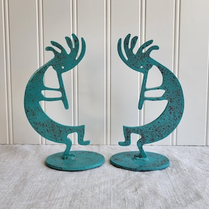 May include: Two turquoise metal Kokopelli sculptures with a distressed finish. Each sculpture features a stylized figure with a curved body, playing a flute, and stands on a round base. The sculptures are approximately 6 inches tall.
