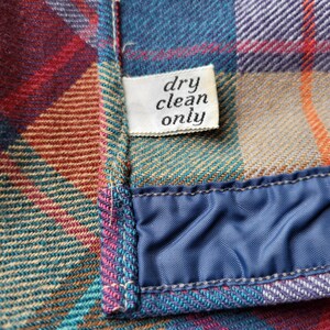Peut inclure: Gros plan sur un tissu &agrave; carreaux dans les tons de bleu sarcelle, rouge, orange et violet. Une petite &eacute;tiquette blanche indique "dry clean only". Le tissu est bord&eacute; d'un mat&eacute;riau bleu satin&eacute;.