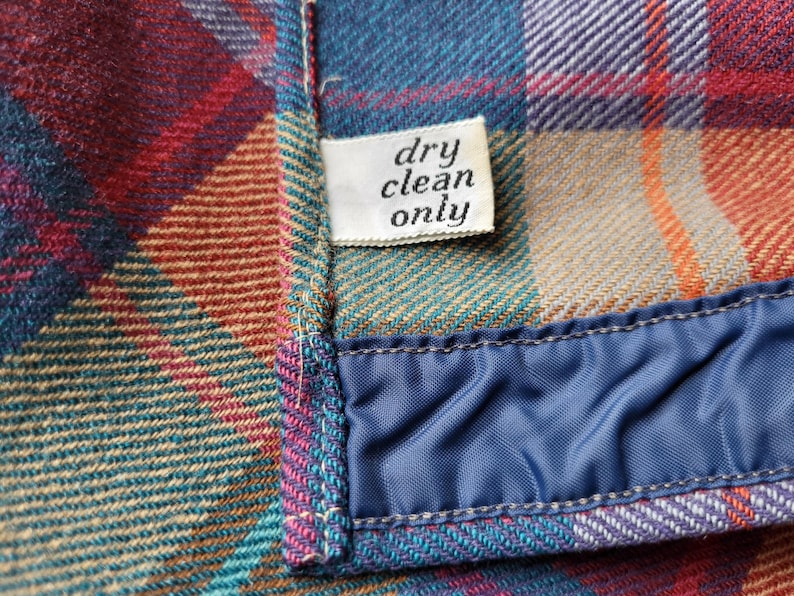 Peut inclure: Gros plan sur un tissu &agrave; carreaux dans les tons de bleu sarcelle, rouge, orange et violet. Une petite &eacute;tiquette blanche indique "dry clean only". Le tissu est bord&eacute; d'un mat&eacute;riau bleu satin&eacute;.