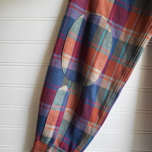 Peut inclure: Gros plan sur une manche de chemise &agrave; carreaux avec un coudi&egrave;re. La chemise pr&eacute;sente un motif de carr&eacute;s bleus, orange, rouges et verts. Le coudi&egrave;re est de couleur beige uni. Le poignet a une fermeture &agrave; bouton.