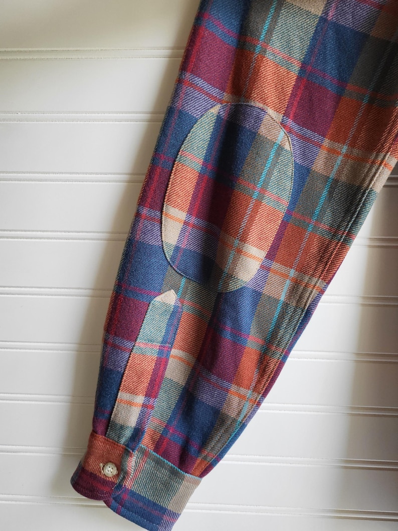 Peut inclure: Gros plan sur une manche de chemise &agrave; carreaux avec un coudi&egrave;re. La chemise pr&eacute;sente un motif de carr&eacute;s bleus, orange, rouges et verts. Le coudi&egrave;re est de couleur beige uni. Le poignet a une fermeture &agrave; bouton.