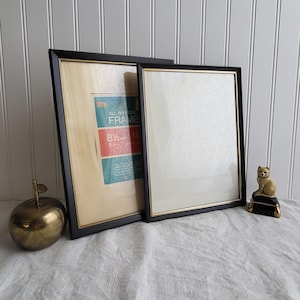 May include: Two black picture frames with gold trim, one with a label that reads "ALL PURPOSE FRAME 8 1/2 x 11". A brass apple and a small cat figurine are also present. The frames are leaning against a white paneled wall.
