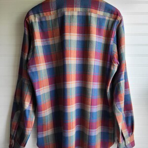 Peut inclure: Une chemise &agrave; carreaux &agrave; manches longues dans des tons de bleu, orange, rouge et beige. La chemise est suspendue &agrave; un cintre en bois. Elle pr&eacute;sente un col boutonn&eacute; et des poignets boutonn&eacute;s. Le fond est blanc.