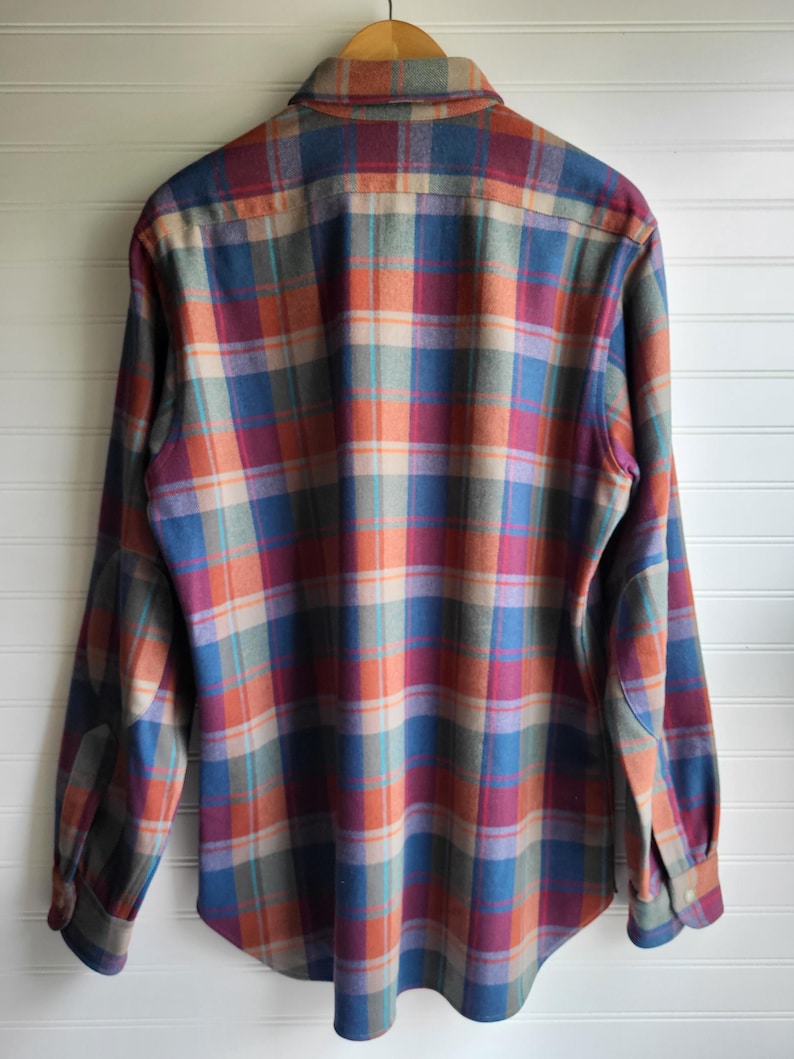 Peut inclure: Une chemise &agrave; carreaux &agrave; manches longues dans des tons de bleu, orange, rouge et beige. La chemise est suspendue &agrave; un cintre en bois. Elle pr&eacute;sente un col boutonn&eacute; et des poignets boutonn&eacute;s. Le fond est blanc.