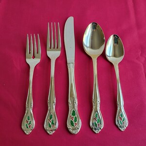 May include: A set of five silver-colored flatware pieces arranged on a red surface. The set includes two forks, a knife, and two spoons. The handles feature a green holly design with red berries, adding a festive touch to the silverware.
