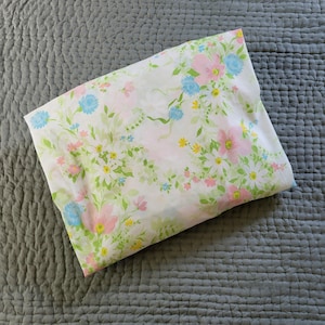 May include: A rectangular fabric item featuring a white background with a repeating floral pattern. The design includes pink, light blue, and yellow flowers interspersed with green leaves and stems, creating a soft, botanical aesthetic across the surface.