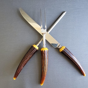 May include: A set of three serving utensils: a carving knife, a carving fork, and a sharpening steel. The handles are brown with yellow accents. The blades and steel are silver.