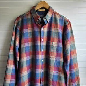 May include: A button-down plaid shirt in a variety of colors, including blue, red, orange, and green. The shirt has a button-down collar, a chest pocket, and long sleeves. The shirt is hanging on a wooden hanger.