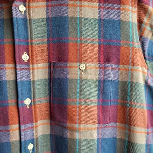 Peut inclure: Gros plan d'une chemise boutonn&eacute;e &agrave; carreaux bleus, oranges, verts et bordeaux. La chemise a une poche poitrine et des boutons blancs. Le tissu semble &ecirc;tre une mati&egrave;re douce et tiss&eacute;e.