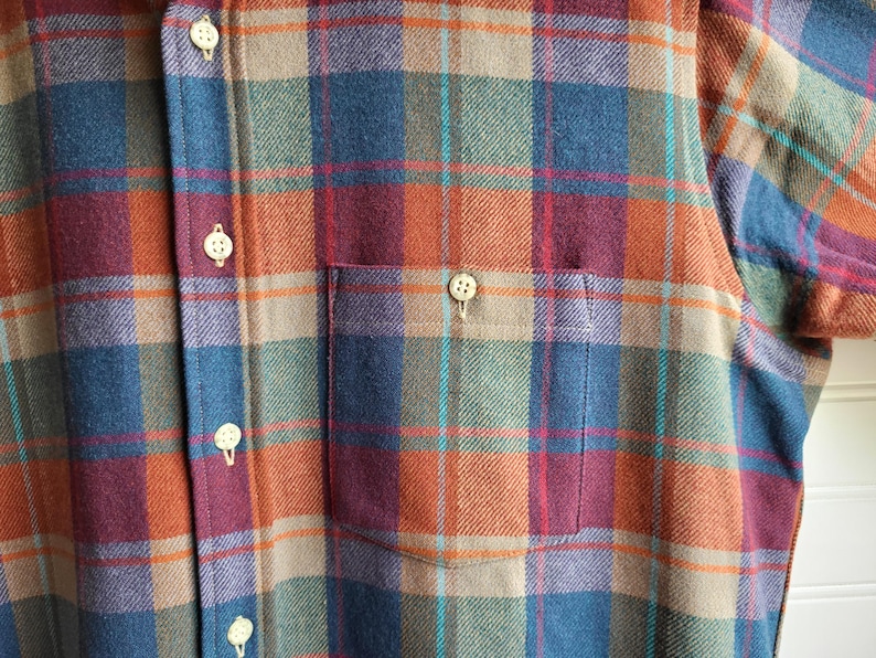 Peut inclure: Gros plan d'une chemise boutonn&eacute;e &agrave; carreaux bleus, oranges, verts et bordeaux. La chemise a une poche poitrine et des boutons blancs. Le tissu semble &ecirc;tre une mati&egrave;re douce et tiss&eacute;e.
