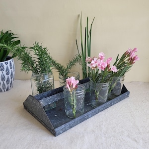 May include: A galvanized metal tray holds several small glass jars filled with greenery and pink flowers. Rosemary sprigs and tall green grass are in some jars. The jars are arranged on a rectangular tray, creating a rustic, natural display.