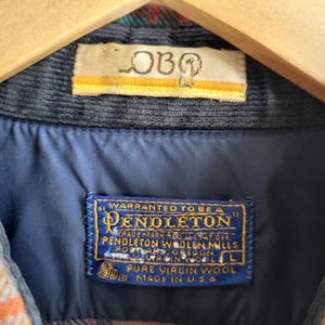 Peut inclure: Gros plan sur une chemise en laine Pendleton avec un col en velours c&ocirc;tel&eacute; bleu et une doublure bleue. La chemise pr&eacute;sente un motif &agrave; carreaux color&eacute; orange, sarcelle et rouge. L'&eacute;tiquette Pendleton est visible, avec le texte "Pure Virgin Wool Made in U.S.A."