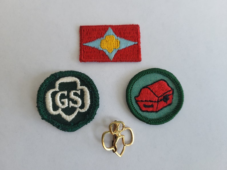 May include: Four Girl Scout badges, including a red and blue badge with a yellow star, a green badge with the letters GS, a green badge with a red treasure chest, and a gold pin with a girl scout silhouette.