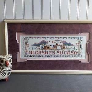 May include: A framed cross-stitch with a burgundy mat and a white background. The cross-stitch depicts a scene of adobe houses with the text "Mi Casa Es Su Casa".