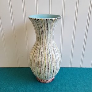 May include: A tall, decorative vase with a flared top and a bulbous base. The vase features a textured surface with vertical stripes in various colors, including black, green, yellow, and brown. The interior rim is light blue, and the base has a patterned design.