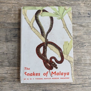 May include: A vintage book cover with a red and black snake wrapped around a branch. The book title is "The Snakes of Malaya" by M.W.F. Tweedie, Raffles Museum, Singapore.