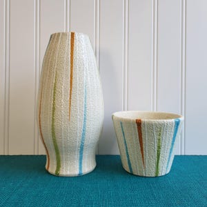 May include: Two ceramic vases with vertical stripes in orange, green, and blue. The taller vase is a slender, elongated shape, while the shorter one is a small, open bowl. Both have a textured, off-white surface.