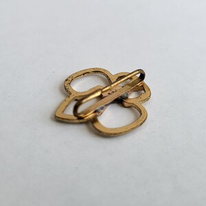 May include: Gold-toned metal clasp with a clover design. The clasp is open and ready to be attached to a garment.