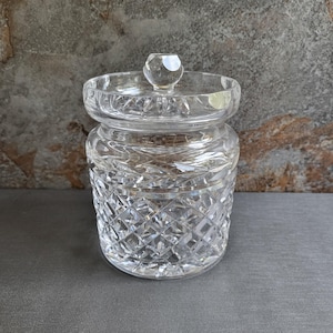 May include: A clear, cut-glass jar with a lid. The jar has a round shape with a diamond pattern on the body and a decorative lid with a small knob. The glass reflects light, creating a sparkling effect. The jar is likely used for storage.