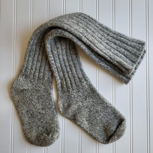 May include: A pair of gray, knee-high socks made of a thick, ribbed knit fabric. The socks are a speckled gray color and appear to be warm and comfortable. The socks are lying flat against a white, slatted background.