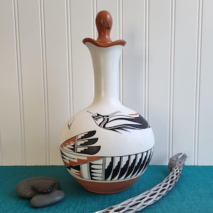 May include: A white ceramic vase with a brown base and lid. The vase features black and orange geometric designs and a stylized bird motif. The lid has a leaf-shaped detail. The vase is approximately 12 inches tall.