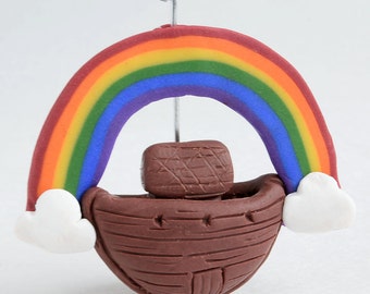 May include: A handcrafted ornament featuring a brown ark with a rainbow arching over it. The rainbow displays the colors red, orange, yellow, green, blue, and purple. White clouds are on either side of the ark. A silver wire is attached for hanging.
