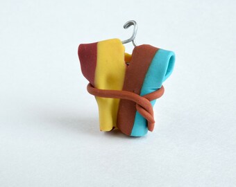 May include: A miniature clay sculpture of a garment on a wire hanger. The garment is composed of yellow, red, and blue clay, with a brown clay belt wrapped around the middle. The background is plain white.