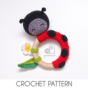 May include: A crocheted ladybug rattle toy with a wooden ring, red and black crochet body, gray face, and a green leaf and white flower attached.