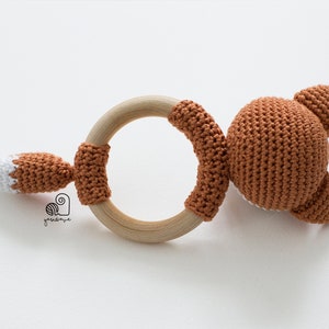 May include: A crocheted fox rattle toy with a wooden ring. The fox is brown with a white tail tip.