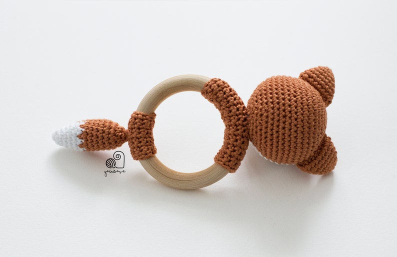 May include: A crocheted fox rattle toy with a wooden ring. The fox is brown with a white tail tip.