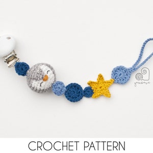 Könnte beinhalten: Gehäkelter Schnullerhalter für Babys mit einem grau-weißen Pinguin, einem gelben Stern und blauen Kugeln. Der Clip hat einen weißen Holzknopf und einen Metallclip. Der Text "CROCHET PATTERN" steht unten.