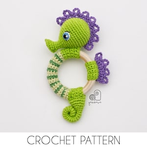 Może przedstawiać: Zielona, dziana grzechotka w kształcie konika morskiego z drewnianym pierścieniem. Konik morski ma fioletowe, dziane detale i niebieskie oko.