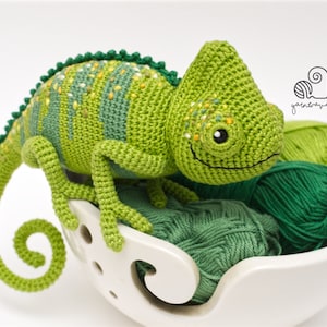 Puede incluir: Un juguete de camale&oacute;n de crochet verde con manchas amarillas, sentado en un cuenco de lana blanco con ovillos de lana verdes. El juguete tiene una cara sonriente y una cola enroscada.